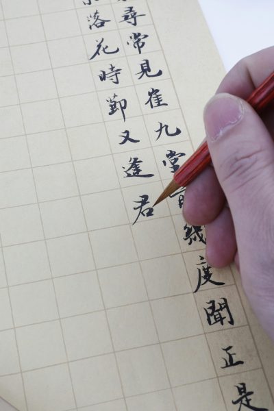 Detail of a hand writing traditional Chinese calligraphy with a brush on grid-lined paper.