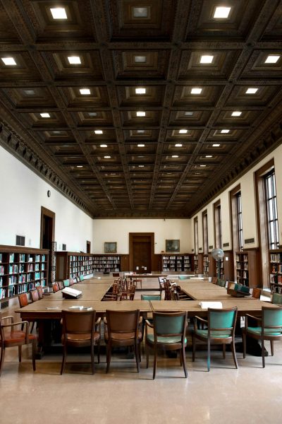 Spacious library with wooden tables and chairs, surrounded by bookcases, illuminated by ceiling lights.